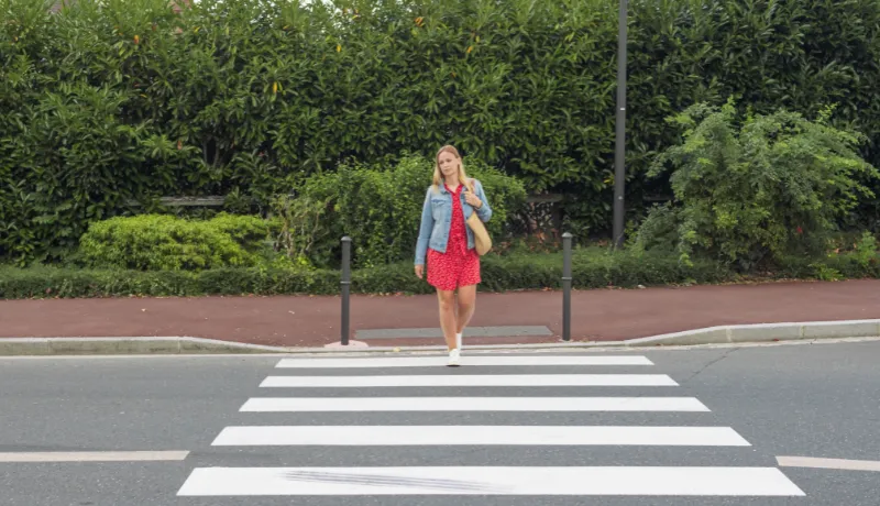 A girl with fair hair in a red dress and a denim jacket with a bag on her shoulder crosses the road at a pedestrian crossing.