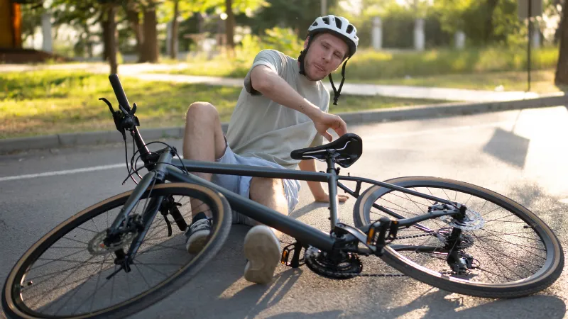 man wearing a helmet sits on the asphalt of a city street, his leg injured, after a bicycle crash