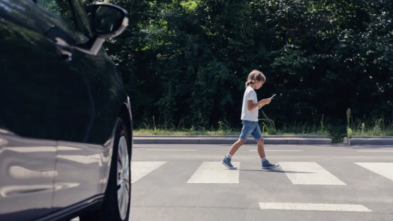Low angle of car in front of pedestrian crossing and walking boy with smartphone