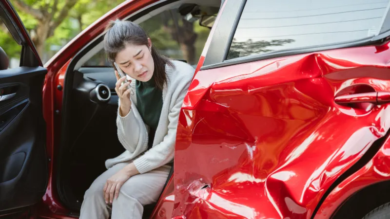 A woman is sitting in a red car with a smashed door. She is talking on her cell phone.