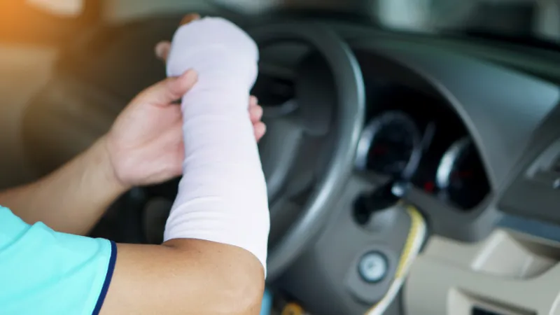 Injured man with bandage on hand and arm is driving car that might cause accident because it's uncomfortable to drive as normal hand.