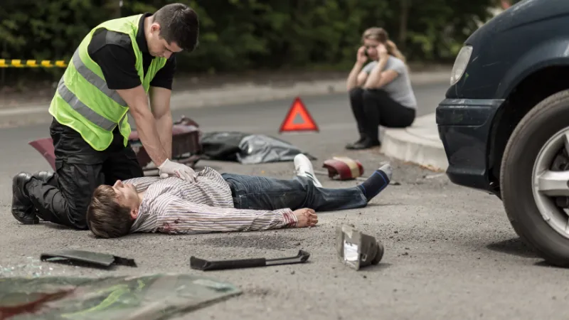 Paramedic resuscitating on street car accident victim, in the background woman talking on phone.